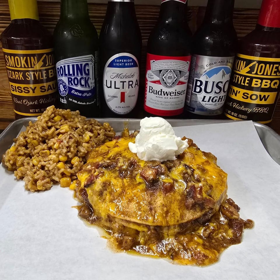A plate of cheesy chili mac with a side of rice and corn, with beer bottles in the background.