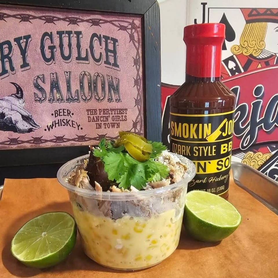 A plastic cup of elote with meat, cilantro, and jalapeños, next to limes and BBQ sauce.