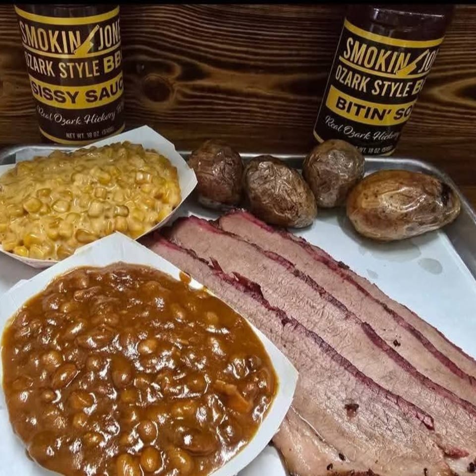 A tray of sliced brisket, baked beans, creamed corn, and baked potatoes with two BBQ sauce bottles.