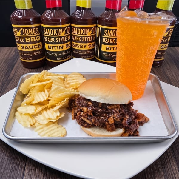 A BBQ pulled pork sandwich with chips and an orange soda, with Smokin' Jones BBQ sauces in the background.