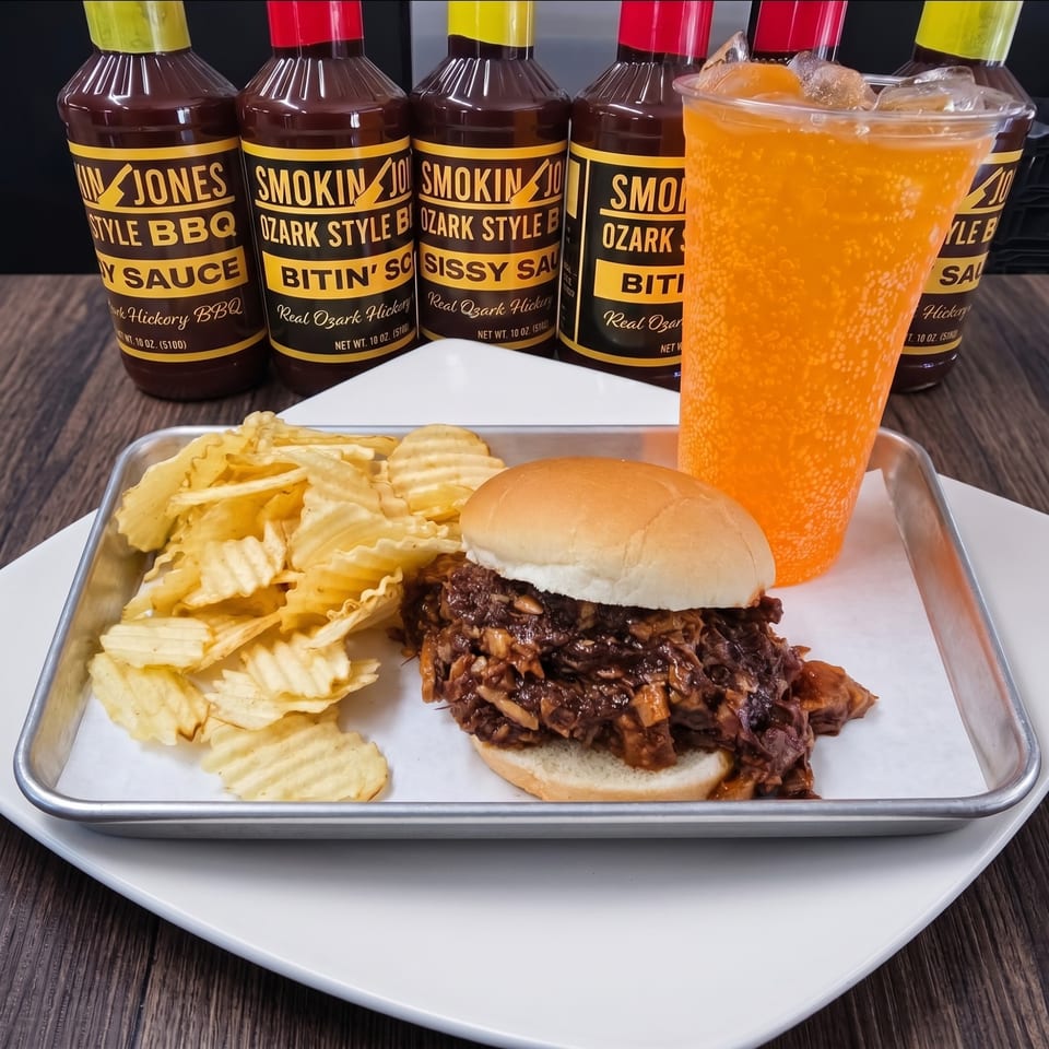 A BBQ pulled pork sandwich with chips and an orange soda, with Smokin' Jones BBQ sauces in the background.