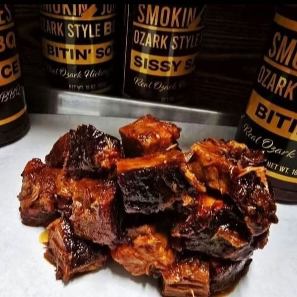 Pile of glistening, dark brown BBQ burnt ends with three bottles of "Smokin' Ozark Style" BBQ sauce in the background.