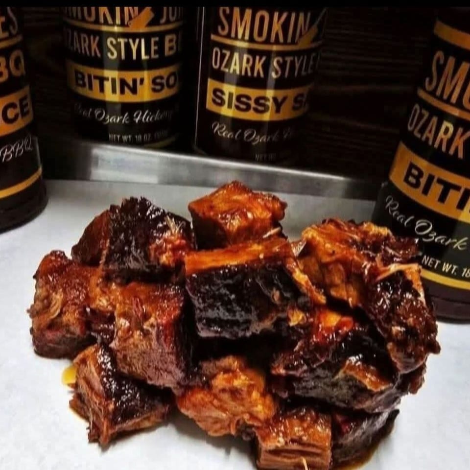 Pile of glistening, dark brown BBQ burnt ends with three bottles of "Smokin' Ozark Style" BBQ sauce in the background.