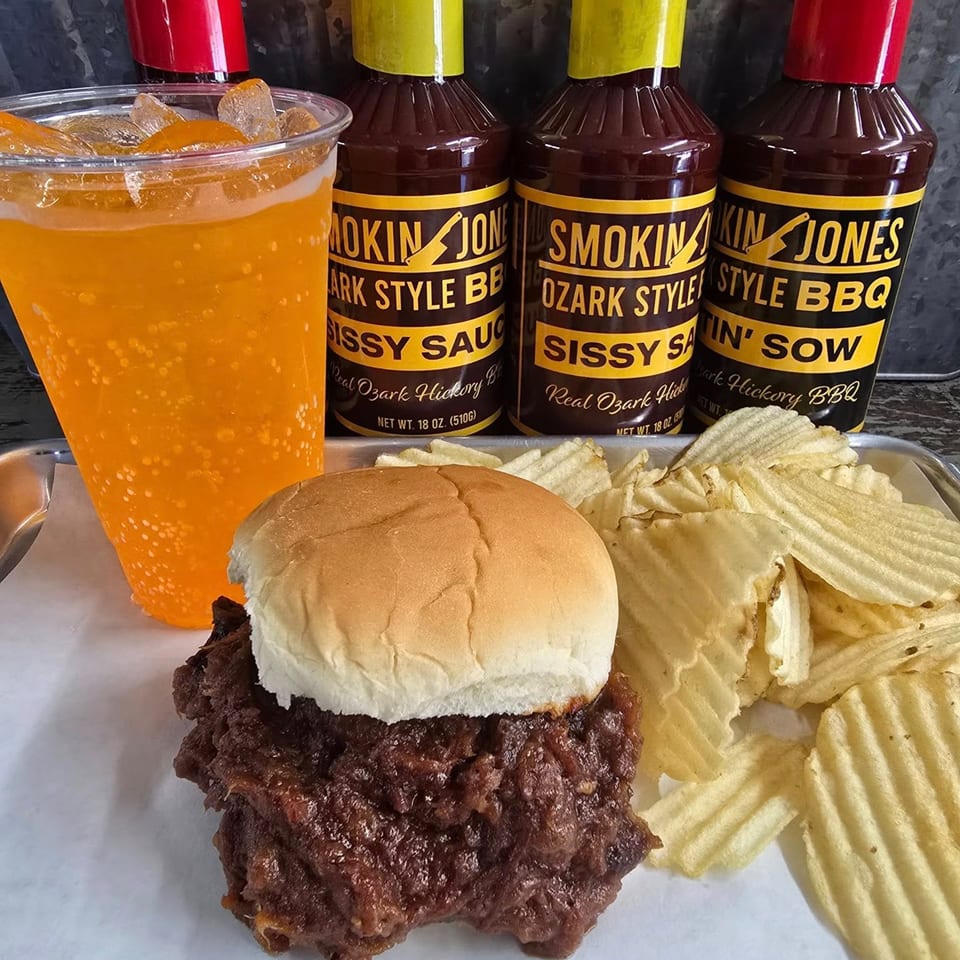 Pulled pork sandwich with chips, an orange drink, and three bottles of Smokin' Jones BBQ sauce.